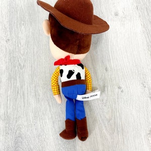 May include: A plush toy of Woody from Toy Story, wearing his signature brown cowboy hat, red bandana, and blue jeans. The toy has a black and white cow pattern on its shirt and a Disney Pixar tag.