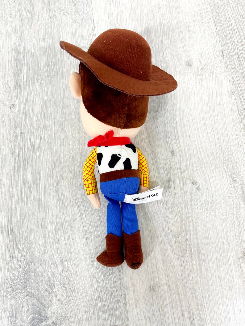 May include: A plush toy of Woody from Toy Story, wearing his signature brown cowboy hat, red bandana, and blue jeans. The toy has a black and white cow pattern on its shirt and a Disney Pixar tag.