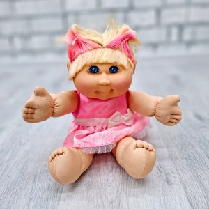 Puede incluir: Muñeca Cabbage Patch Kids con cabello rubio y rosa, ojos azules y un vestido rosa. La muñeca está sentada con los brazos extendidos. El vestido tiene un ribete de encaje blanco y un lazo blanco.