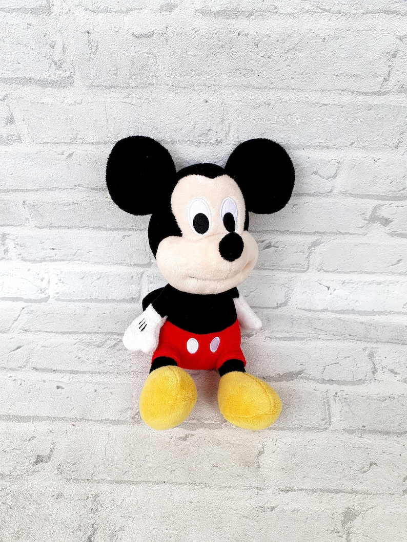 May include: A plush Mickey Mouse toy with black ears, a black nose, and white eyes. The toy is wearing red shorts with white polka dots and yellow shoes.