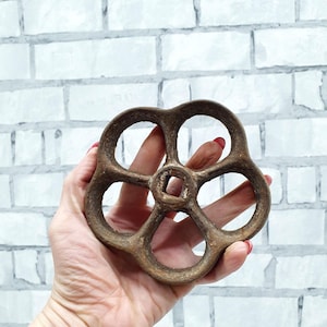 May include: A vintage, bronze-colored valve wheel with a six-spoke design. The wheel has a central square opening and is held against a white brick wall. The wheel's aged appearance suggests it is an antique or reclaimed industrial piece.