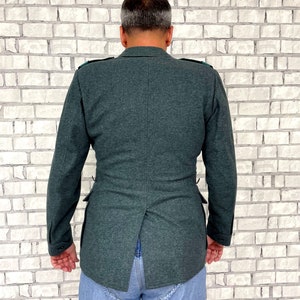 May include: A dark green jacket with a back vent and a single button closure. The jacket has a textured fabric and is worn over a pair of blue jeans.