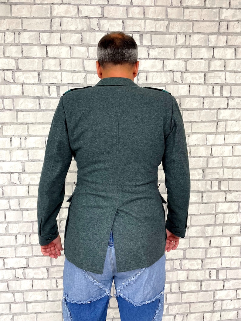 May include: A dark green jacket with a back vent and a single button closure. The jacket has a textured fabric and is worn over a pair of blue jeans.