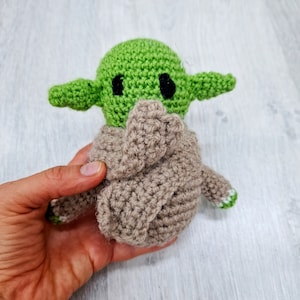 May include: A green and brown crocheted plush toy resembling a popular fictional character. The toy has large eyes and a small mouth.