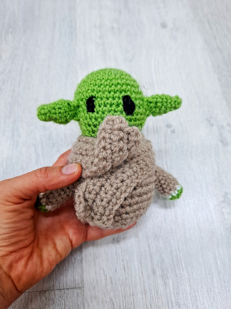 May include: A green and brown crocheted plush toy resembling a popular fictional character. The toy has large eyes and a small mouth.