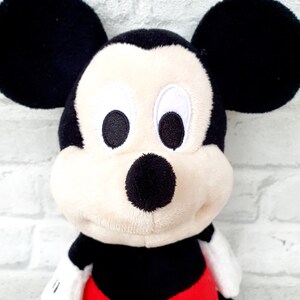 May include: A plush Mickey Mouse toy with black ears, a black nose, and red shorts with white polka dots. The toy has a white face with black eyes and a big smile.