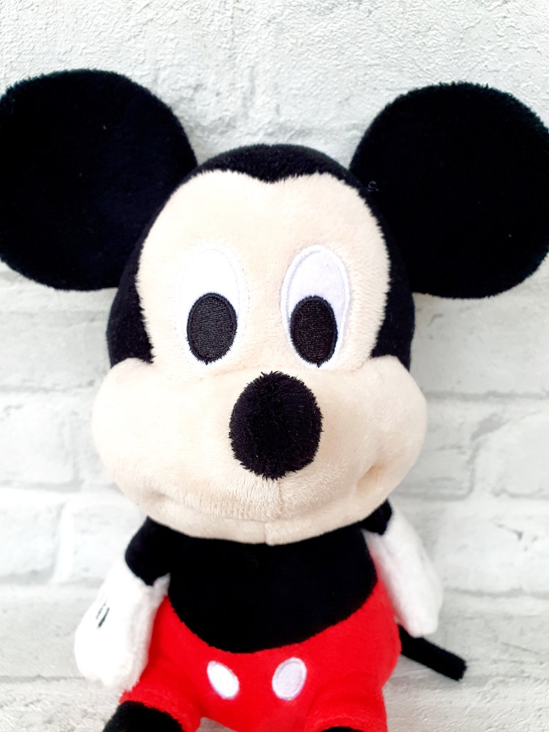 May include: A plush Mickey Mouse toy with black ears, a black nose, and red shorts with white polka dots. The toy has a white face with black eyes and a big smile.