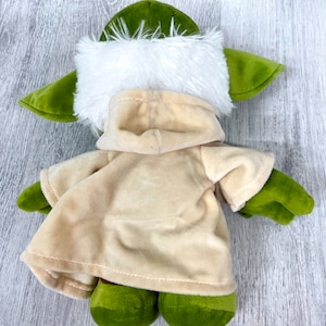 May include: Green plush toy with a white fur hood and a tan robe. The toy is lying on a wooden surface.