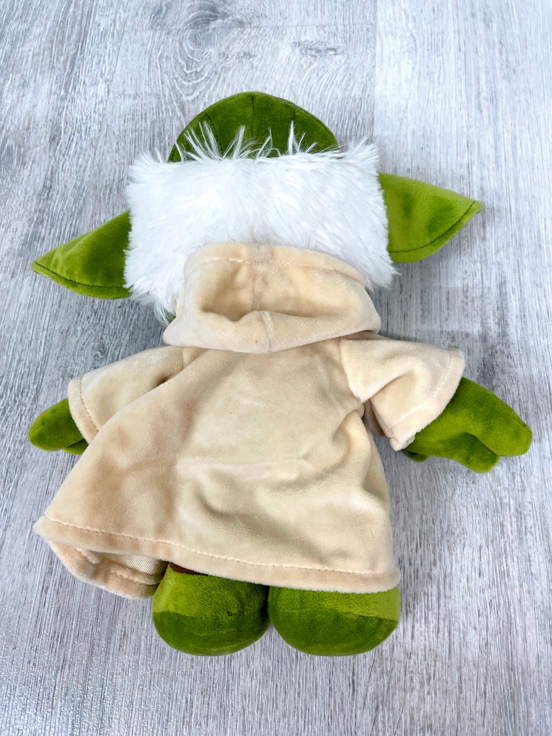May include: Green plush toy with a white fur hood and a tan robe. The toy is lying on a wooden surface.
