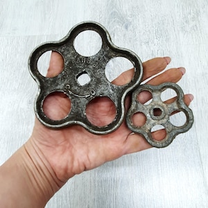 May include: Two antique, cast-iron valve handles. The larger handle has a five-petal design with circular openings and the text "AMHO CO." The smaller handle has a similar design. Both are held in a hand.