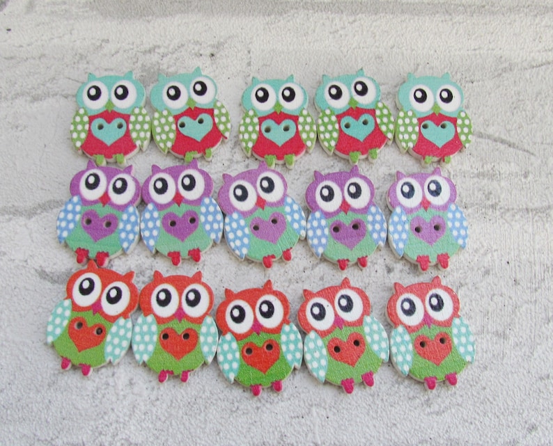 May include: A set of 15 wooden owl buttons with colorful designs. The owls have big eyes, a heart shape on their chest, and polka dots on their wings. The buttons are in shades of green, red, blue, purple, and white.
