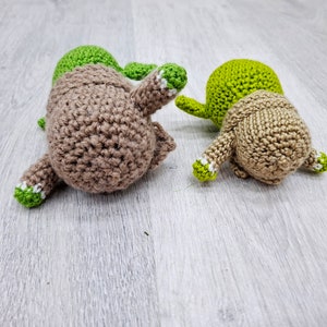 May include: Two crocheted stuffed toys, one brown and one green, lying on a light grey wood surface. Both toys have white accents on their limbs.
