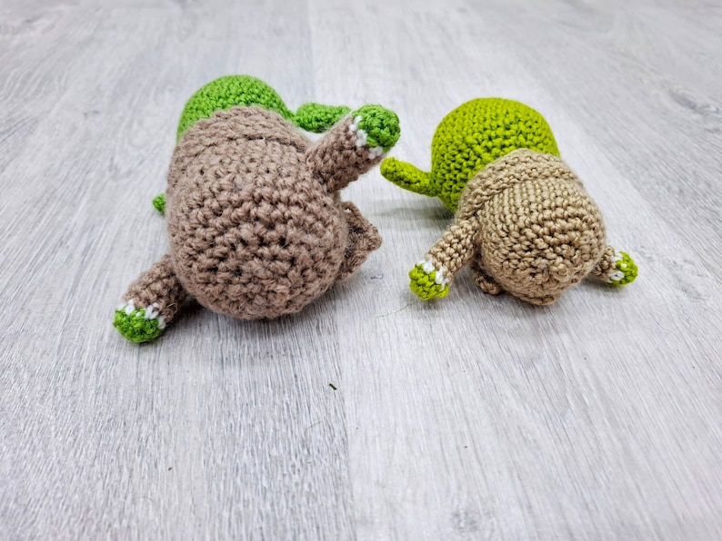 May include: Two crocheted stuffed toys, one brown and one green, lying on a light grey wood surface. Both toys have white accents on their limbs.
