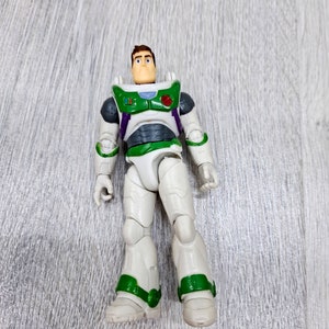 Puede incluir: Una figura de acci&oacute;n de pl&aacute;stico blanco y verde de Buzz Lightyear de la franquicia Toy Story. La figura tiene el pelo marr&oacute;n y un traje espacial verde y blanco con un bot&oacute;n rojo en el pecho.