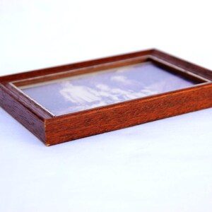 Puede incluir: Un marco de fotos rectangular de madera con acabado marr&oacute;n oscuro. El marco contiene una fotograf&iacute;a en blanco y negro bajo vidrio. El marco est&aacute; plano sobre una superficie blanca.