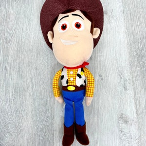 May include: A plush toy of Woody from Toy Story. He is wearing his signature brown cowboy hat, red bandana, yellow and red plaid shirt, blue jeans, and brown boots. He has a friendly smile and brown eyes.