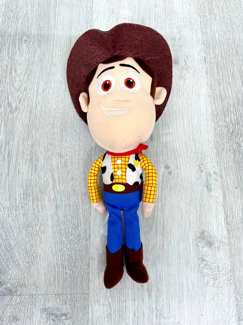 May include: A plush toy of Woody from Toy Story. He is wearing his signature brown cowboy hat, red bandana, yellow and red plaid shirt, blue jeans, and brown boots. He has a friendly smile and brown eyes.