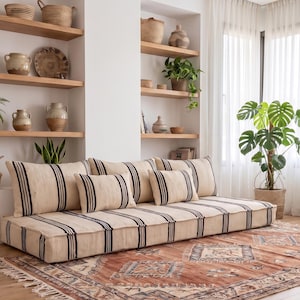 May include: A low-profile floor sofa featuring a cream-colored fabric with vertical black stripes, accompanied by matching back cushions and throw pillows. The sofa is positioned on a large, rectangular area rug with a rust-orange and blue pattern.