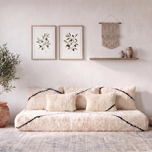 May include: A low-profile, cream-colored faux fur sofa with black line accents, two matching throw pillows, and a large area rug with a faded blue and beige pattern. An olive tree in a terracotta pot stands to the left, with wall art above.