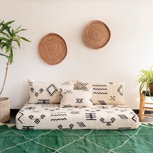 以下が含まれることがあります： 黒の幾何学模様が施された白い床用クッション、黒の幾何学模様が施された枕が2つ、緑の敷物。クッションは白い床に置かれ、壁には籐製のバスケットが2つある。