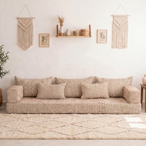 Puede incluir: Un salón de tonos neutros con un sofá grande y bajo y varios cojines. Una alfombra crema con un diseño geométrico está en el suelo. Los tapices de macramé y un olivo en maceta completan la decoración. El sofá mide aproximadamente 200 cm de largo.