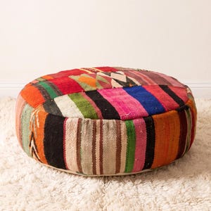 May include: A round floor pouf featuring a patchwork design of woven fabric in vibrant colors including red, orange, green, pink, blue, and black. The textile displays a variety of striped and geometric patterns across the top and sides.