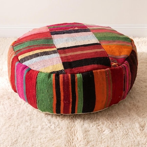 May include: A round floor pouf featuring a patchwork design made from woven fabric strips in vibrant shades of red, green, orange, black, and white. The textile surface displays a variety of horizontal and vertical striped patterns throughout.