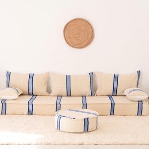 Puede incluir: Un sofá de suelo color crema con rayas azules, acompañado de cojines a juego y un otomán redondo. Un tapiz de pared tejido y pequeñas mesas auxiliares con decoración completan el ambiente de salón de estilo bohemio.