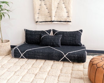 May include: A black and white patterned floor cushion with three black pillows. The cushion is on a white shag rug and there is a brown leather pouf in the foreground.