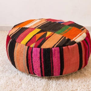 May include: A round floor pouf crafted from woven fabric featuring a patchwork design of multicolored stripes and geometric patterns in shades of red, orange, yellow, pink, and black. The textile surface displays a textured, hand-woven appearance.