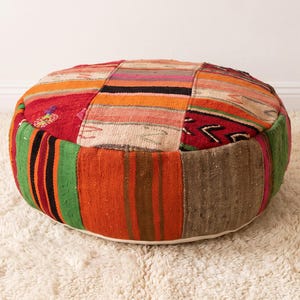 May include: A round floor pouf constructed from various woven textile panels featuring a vibrant patchwork of red, orange, green, brown, and cream colors with geometric patterns and striped designs across the top and sides of the cushion.