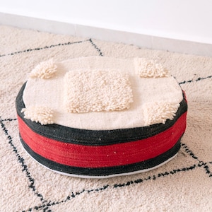 May include: Round, cream-colored pouf with a central square and four corner accents of textured, fluffy material. The sides feature a black band with a red stripe, resting on a patterned rug.