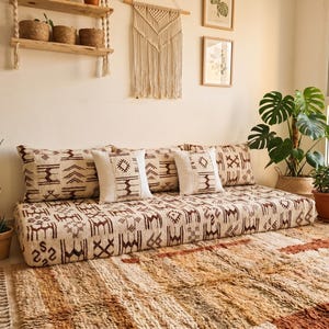 May include: A low-profile floor sofa featuring a cream-colored fabric with a repeating dark brown geometric pattern. The sofa includes two large rectangular back cushions and two smaller square accent pillows, all matching the same patterned design.