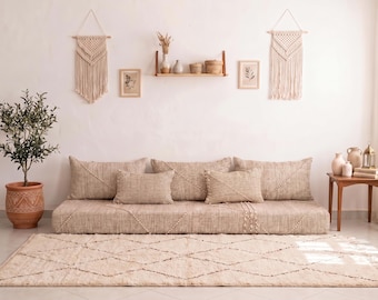 Könnte beinhalten: Ein niedriges beiges Sofa mit strukturierter Stoffoberfläche, ausgestattet mit drei großen Rückenkissen und zwei kleineren rechteckigen Zierkissen. Das Sofa ist mit einem dezenten Rautenmuster versehen und steht auf einem passenden beigen Teppich.