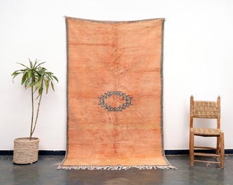 Puede incluir: Una alfombra grande, rectangular, de color naranja con un diseño geométrico azul oscuro en el centro. La alfombra tiene un borde de flecos y está colgada en una pared blanca.