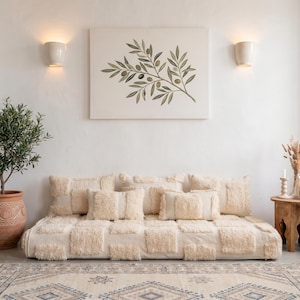 May include: A neutral-toned living room scene featuring a low, tufted sofa with matching pillows. A framed olive branch art piece hangs above. A potted olive tree and a small wooden side table with decor complete the cozy, inviting space.