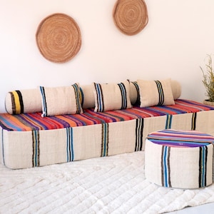 May include: A low sofa with a striped design, accompanied by matching cushions and a round ottoman. The sofa and ottoman have a colourful striped top and vertical stripes on the sides. Woven wall decorations and a small side table are also visible.