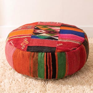 May include: A round floor pouf constructed from various patches of woven fabric featuring a vibrant color palette of red, orange, green, blue, and black. The surface displays a mix of geometric patterns and solid stripes, creating a textured appearance.