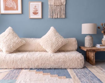 May include: A low-profile cream-colored sofa upholstered in a textured, shaggy fabric. The sofa features a long cylindrical bolster backrest and two matching square throw pillows, all covered in the same plush, high-pile material.