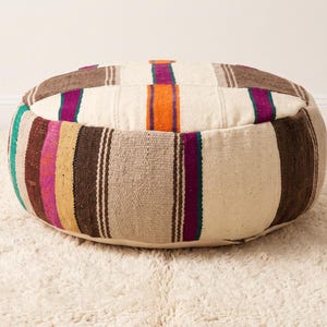 May include: A round floor pouf featuring a woven textile pattern with alternating vertical stripes in cream, dark brown, bright orange, magenta, and teal. The fabric has a visible textured weave, and the design covers the top and sides.