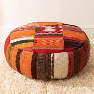 May include: A round floor pouf featuring a patchwork of woven fabric panels in vibrant shades of orange, red, brown, green, and cream. The surface displays a variety of geometric patterns, including stripes, zigzags, and diamonds, with visible stitching.