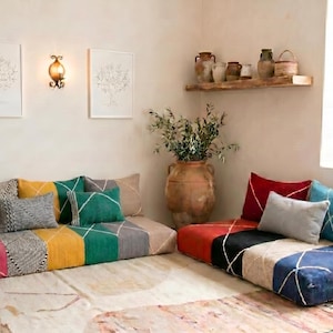 May include: A cozy living space featuring floor cushions in vibrant colors like pink, yellow, green, and blue. Decorative pillows and a large terracotta vase with greenery add to the inviting atmosphere. Artwork and a wall sconce complete the scene.