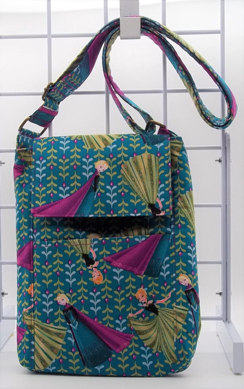 Peut inclure: Un sac &agrave; bandouli&egrave;re bleu sarcelle avec une fermeture &agrave; rabat. Le sac est d&eacute;cor&eacute; d'un motif r&eacute;p&eacute;t&eacute; de personnages de La Reine des Neiges, dont Elsa et Anna, portant leurs robes caract&eacute;ristiques. Le sac a une bandouli&egrave;re rose et violette.