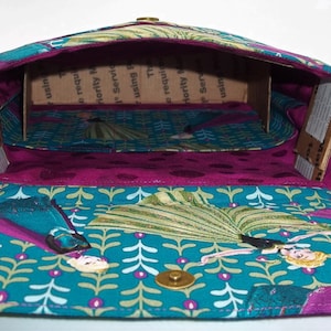 Peut inclure: Un sac en tissu bleu sarcelle avec une doublure violette et un insert en carton brun. Le sac pr&eacute;sente un motif r&eacute;p&eacute;titif de feuilles vertes et de fleurs blanches. Le sac a une boucle en m&eacute;tal dor&eacute; et une fermeture &agrave; pression. Le sac est ouvert et l'int&eacute;rieur est visible.