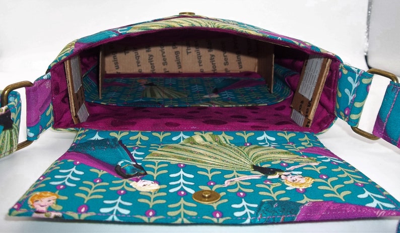 Peut inclure: Un sac en tissu bleu sarcelle avec une doublure violette et un insert en carton brun. Le sac pr&eacute;sente un motif r&eacute;p&eacute;titif de feuilles vertes et de fleurs blanches. Le sac a une boucle en m&eacute;tal dor&eacute; et une fermeture &agrave; pression. Le sac est ouvert et l'int&eacute;rieur est visible.