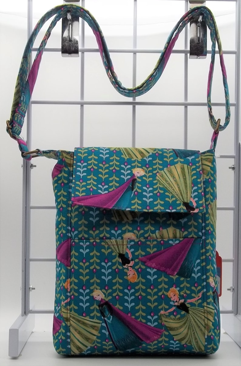 Peut inclure: Un sac &agrave; bandouli&egrave;re bleu sarcelle avec une fermeture &agrave; rabat. Le sac est en tissu avec un motif de feuilles vertes et de fleurs roses. Le sac pr&eacute;sente un imprim&eacute; de personnages du film La Reine des Neiges.