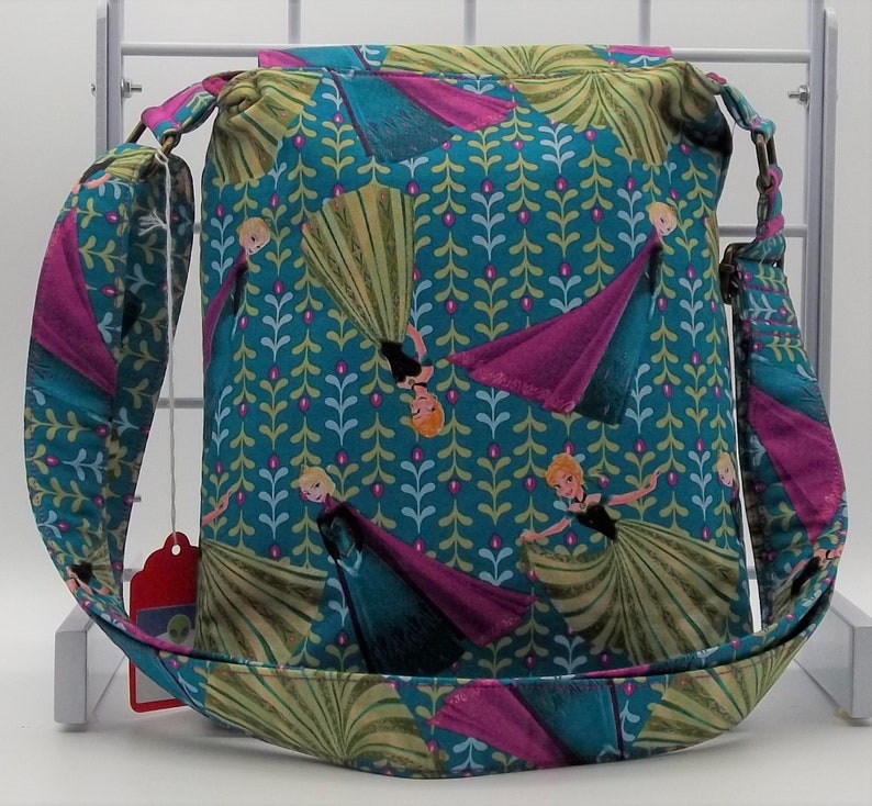 Peut inclure: Un sac en tissu bleu sarcelle avec un motif floral rose et vert repr&eacute;sentant des princesses du film La Reine des Neiges. Le sac a une longue bandouli&egrave;re r&eacute;glable.