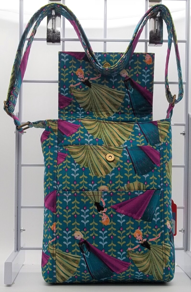 Peut inclure: Un sac &agrave; bandouli&egrave;re bleu sarcelle avec une fermeture &agrave; rabat. Le sac est d&eacute;cor&eacute; d'un motif r&eacute;p&eacute;t&eacute; de feuilles vertes et d'images des personnages du film La Reine des Neiges. Le sac a une fermeture &agrave; bouton dor&eacute;e et une longue bandouli&egrave;re r&eacute;glable.