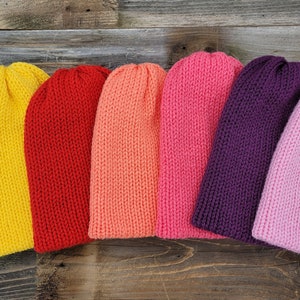 May include: Seven knitted beanies in a rainbow of colors, including green, yellow, red, orange, pink, purple, and light pink. The beanies are arranged in a row on a wooden surface.