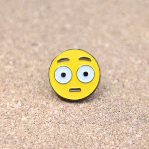 May include: Yellow enamel pin with a cartoon face with wide eyes and a raised eyebrow, resembling a surprised or confused expression.